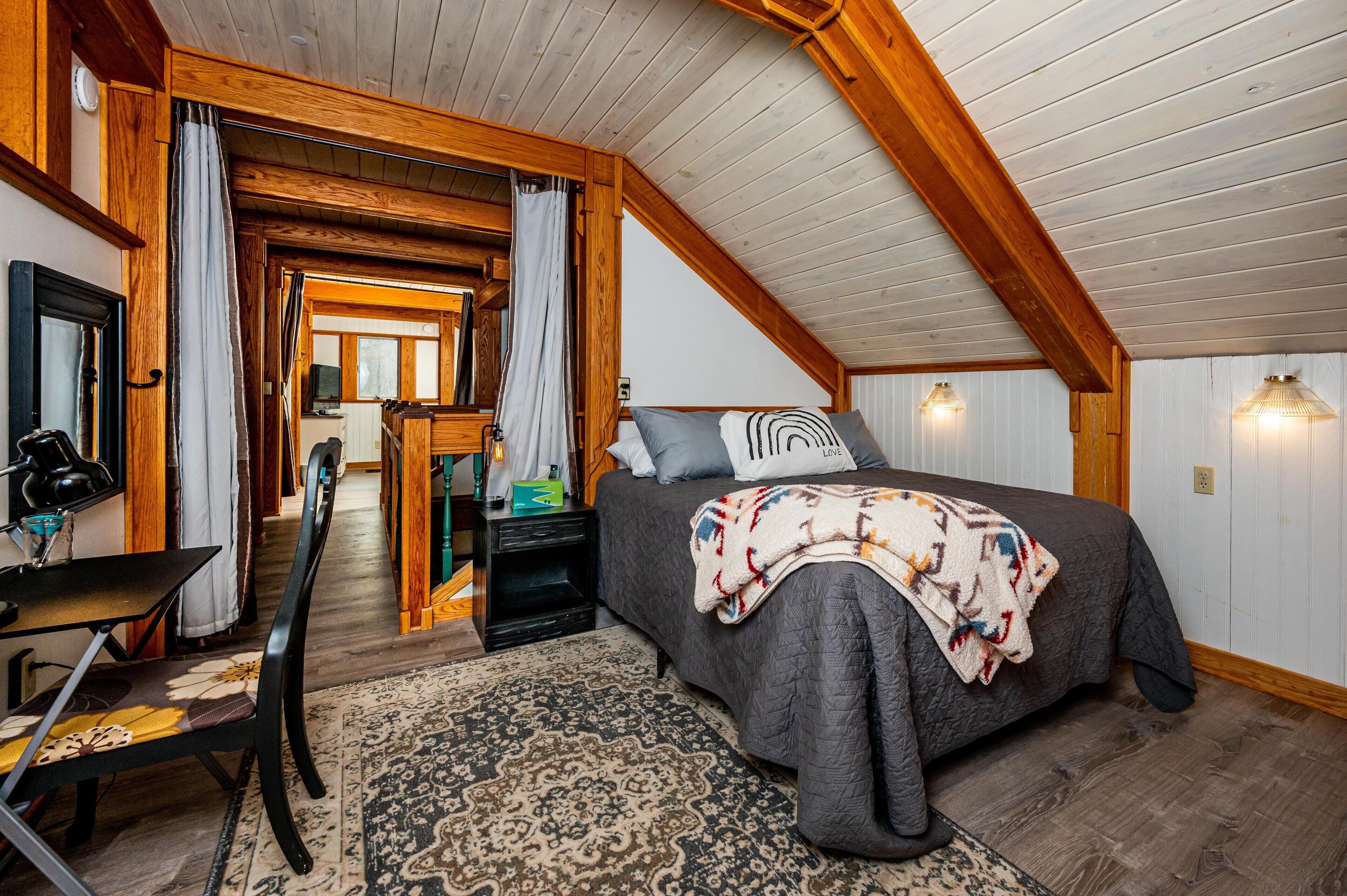 A cozy bedroom with a large bed, a desk, and a chair. There is a wooden ceiling and a window with curtains.