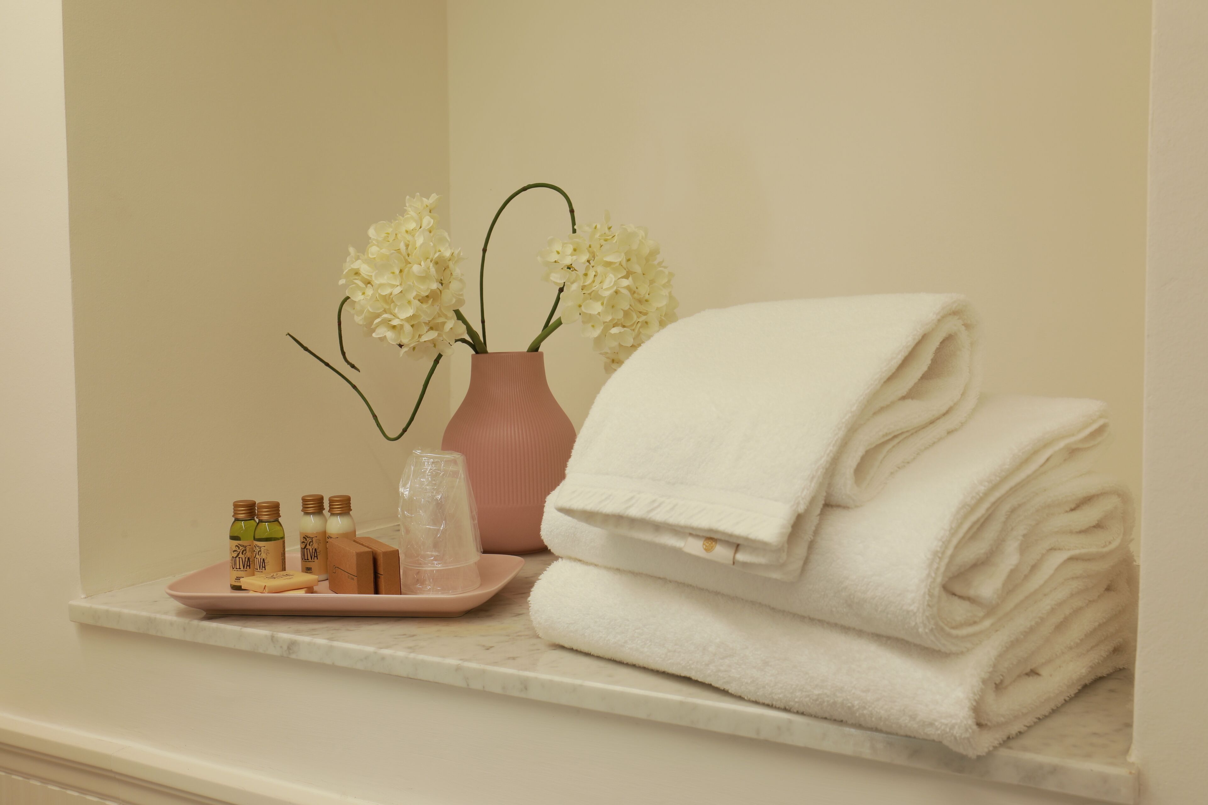 Un tocador de baño con una toalla blanca, un florero rosa con flores blancas y artículos de tocador.