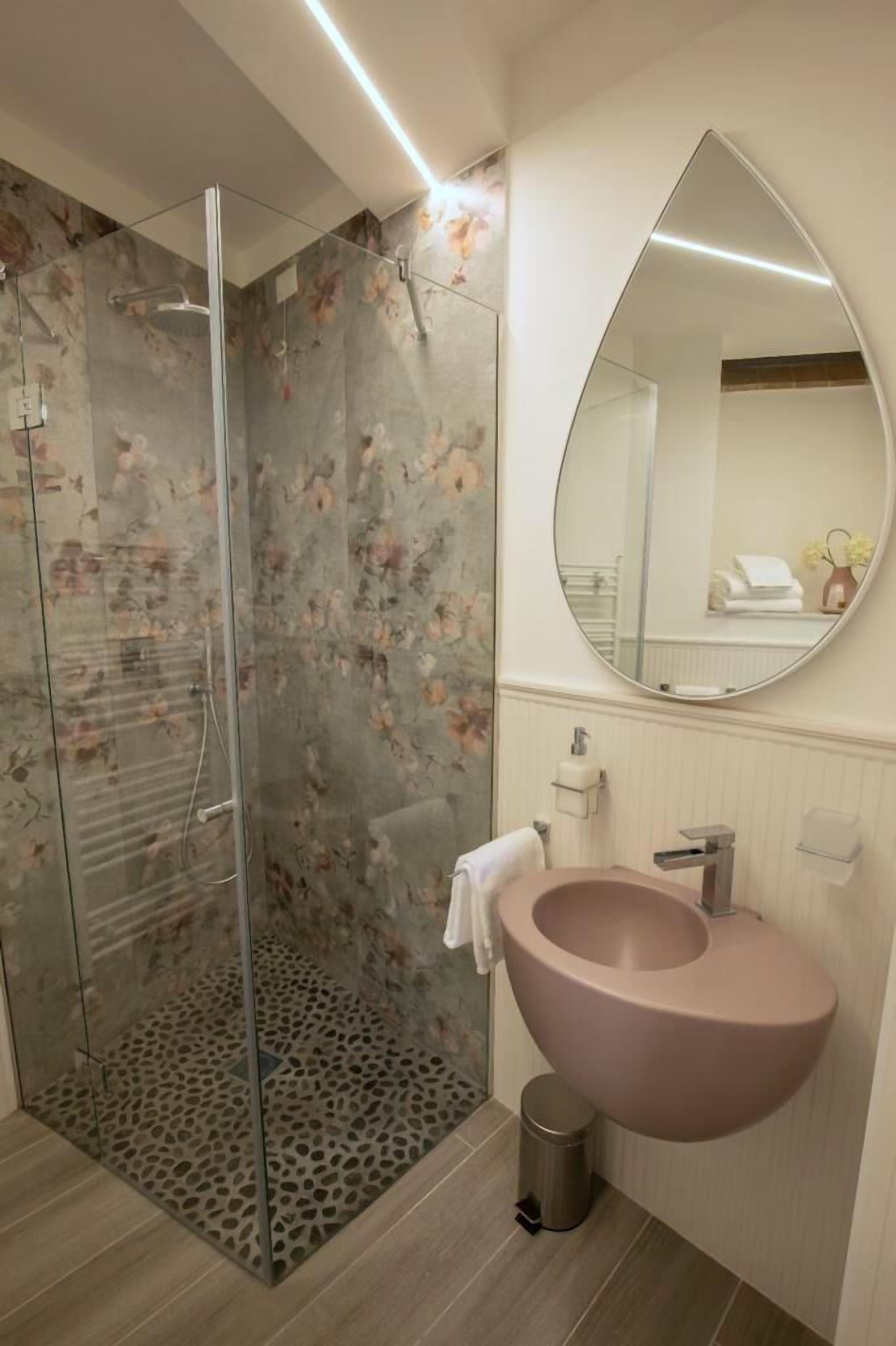 Un baño moderno con cabina de ducha de vidrio, un lavamanos grande de color rosa y un espejo redondo.