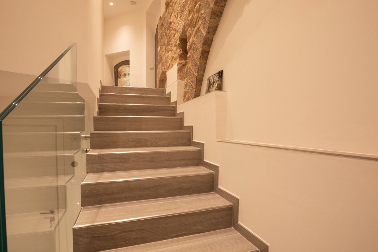 A modern staircase with glass railings and wooden steps, leading to a landing with a stone wall and a framed picture.
