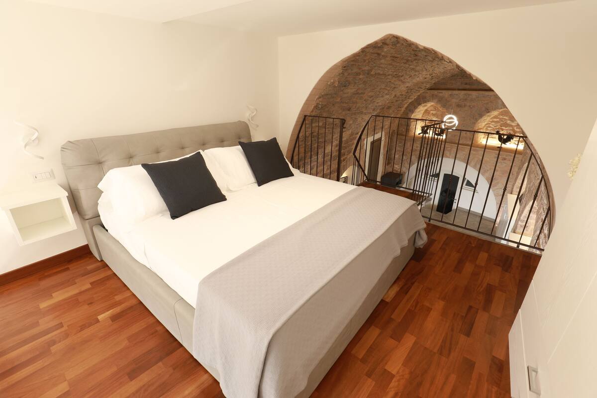 A bedroom with a bed, a wooden floor, a stone archway, and a balcony with a railing.