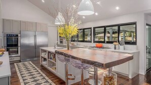A modern kitchen with stainless steel appliances, a large island with bar stools, and a view of the outdoors through large windows. - Calistoga Wine Ranch Estate (Napa Valley)