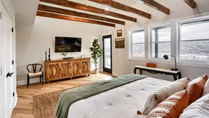 Camera da letto con un letto grande, travi in legno, televisione, una pianta e vista sulla città attraverso ampie finestre.