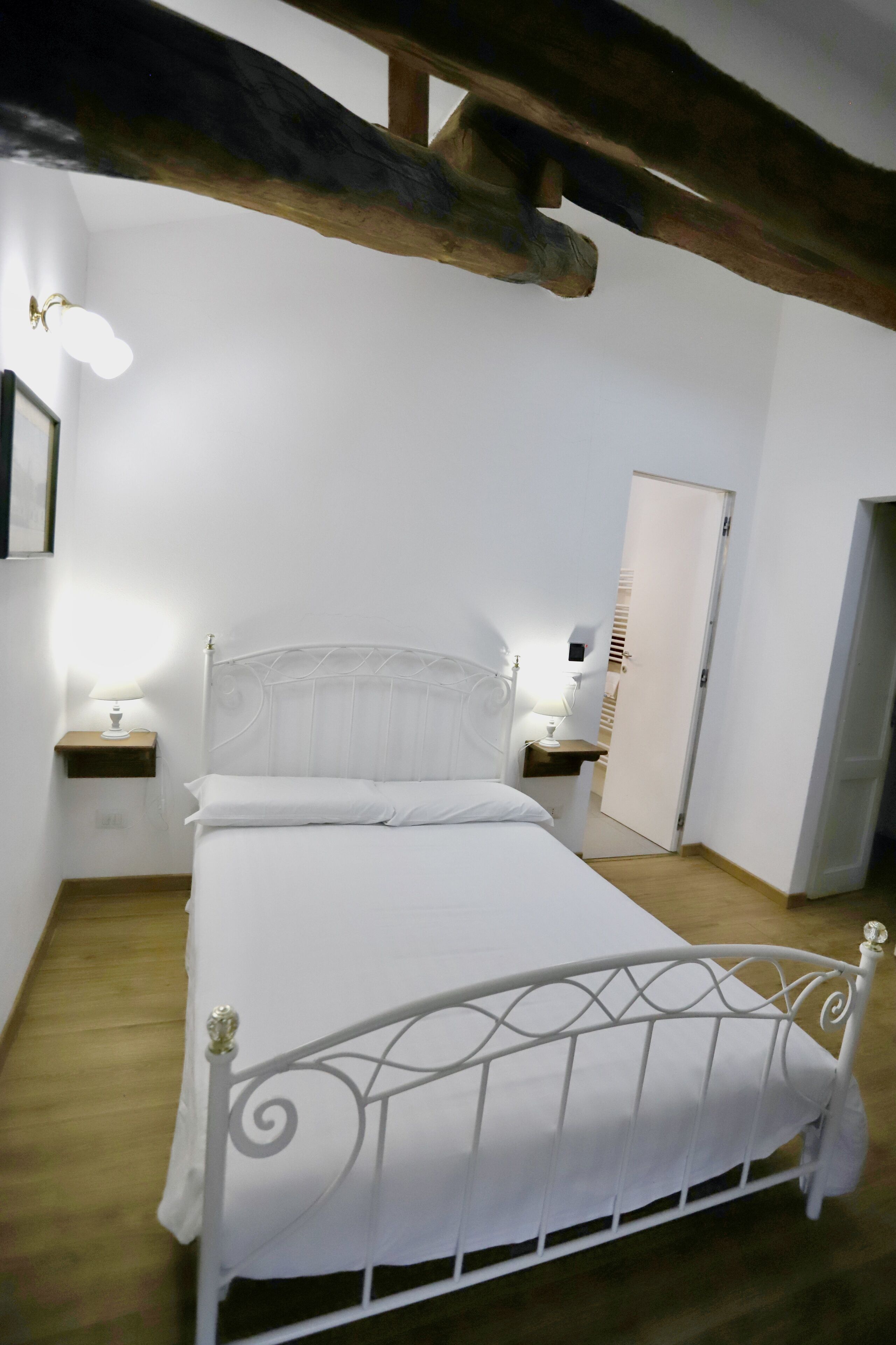 Een slaapkamer met een bed met wit metalen frame, twee nachtkastjes met lampen, een houten balk in het plafond en een witte deur.