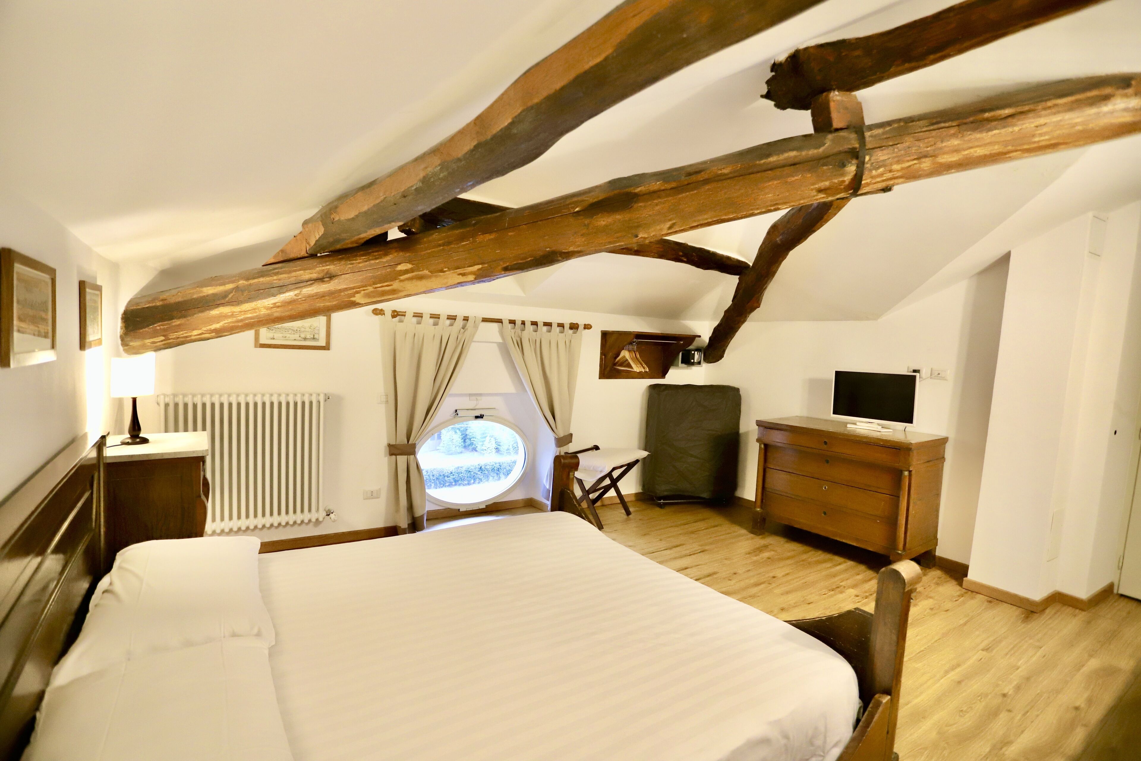 A bedroom with a wooden ceiling, a bed, a dresser, a chair, a desk, a radiator, and a window with curtains.