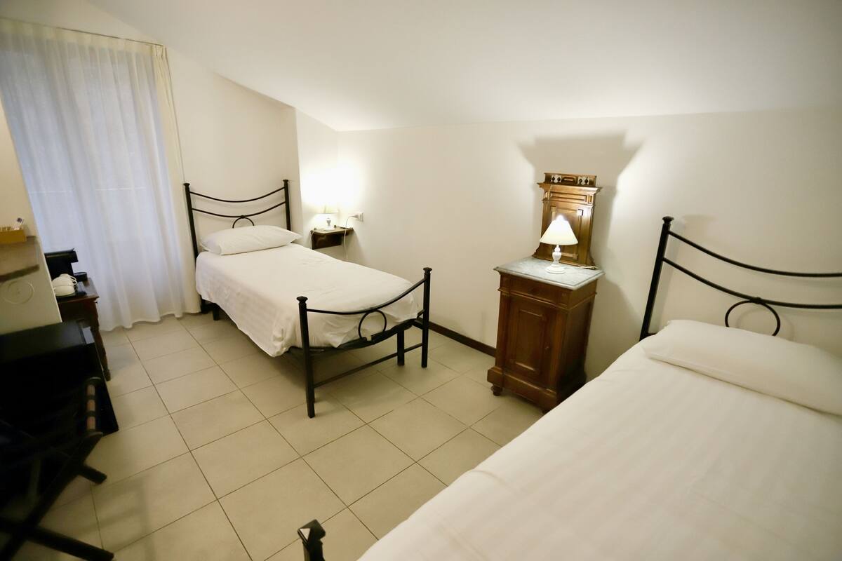 A hotel room with two single beds, a desk with a chair, a nightstand with a lamp, and a wooden cabinet.