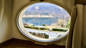 A round window with a view of a lake, trees, and a road. - La Villa Del Lago (Valganna)