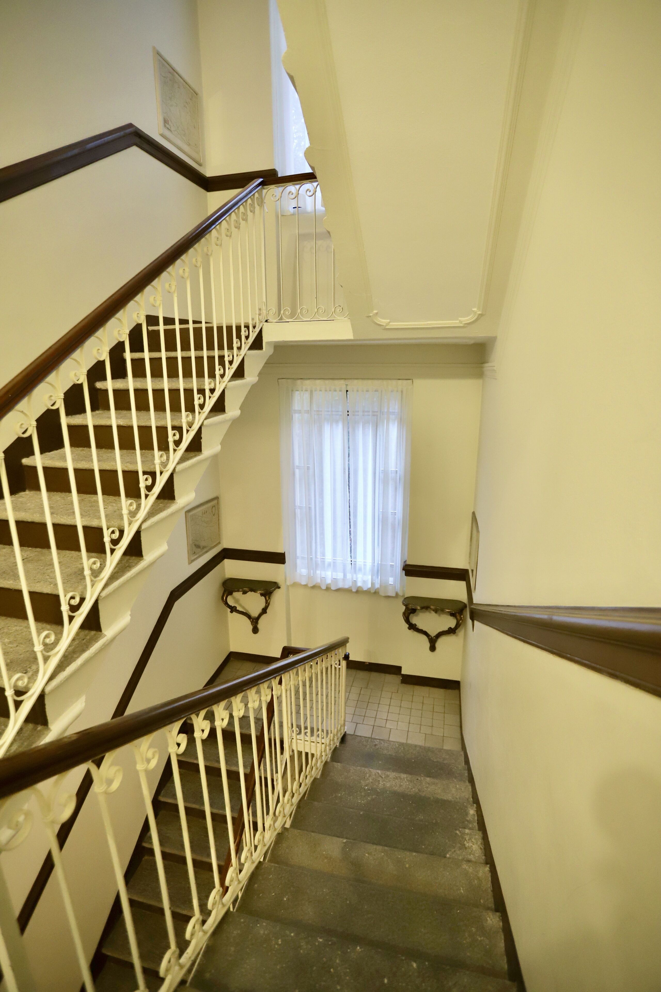 Een trap met witte leuningen en een houten handrail, leidend naar een plateau met een raam met witte gordijnen.