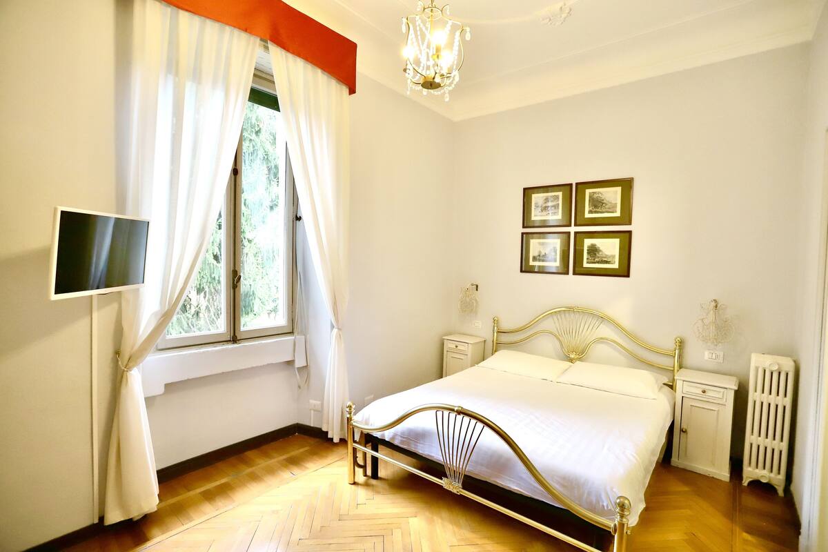 A bedroom with a bed, a TV, a chandelier, framed pictures, and a radiator.