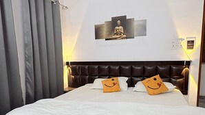Een hotelkamer met een groot bed, grijze gordijnen, een wandairconditioner, een Boeddhabeeld en een kussen met een vrolijk gezicht.