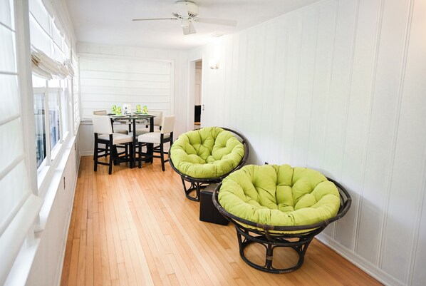 A room with a dining table and chairs, two green cushioned chairs, a ceiling fan, and white paneled walls.