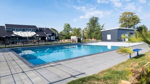 Ein Schwimmbecken mit Fliesen umrandet, ein überdachter Sitzbereich und ein kleines Gebäude im Hintergrund.
