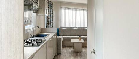Een kleine keuken met een gasfornuis, spoelbak en moderne kasten. Een ingebouwde bank met kussen staat bij een raam met jaloezieën.