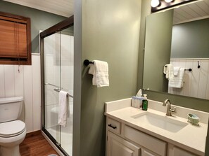 Un baño con cabina de ducha de vidrio, inodoro blanco, lavamanos con espejo y toallero con varias toallas.