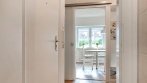 Ein Flur, der in einen Essbereich führt, mit einem weißen Tisch und Stühlen, einem Fenster mit Blick auf Grünflächen und einem Heizkörper unter dem Fenster.