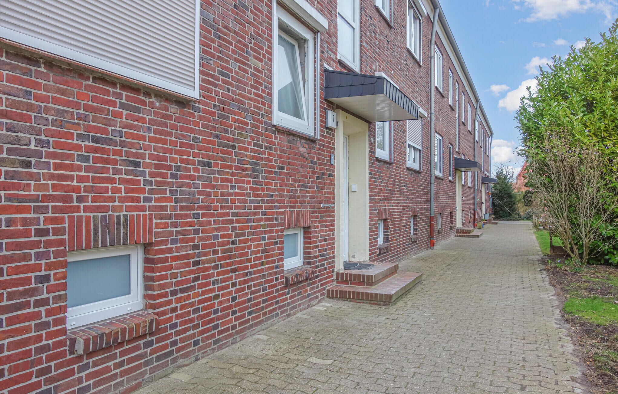 A row of red brick townhouses with white window frames, a paved walkway, and a small garden area with a tree and shrubs.