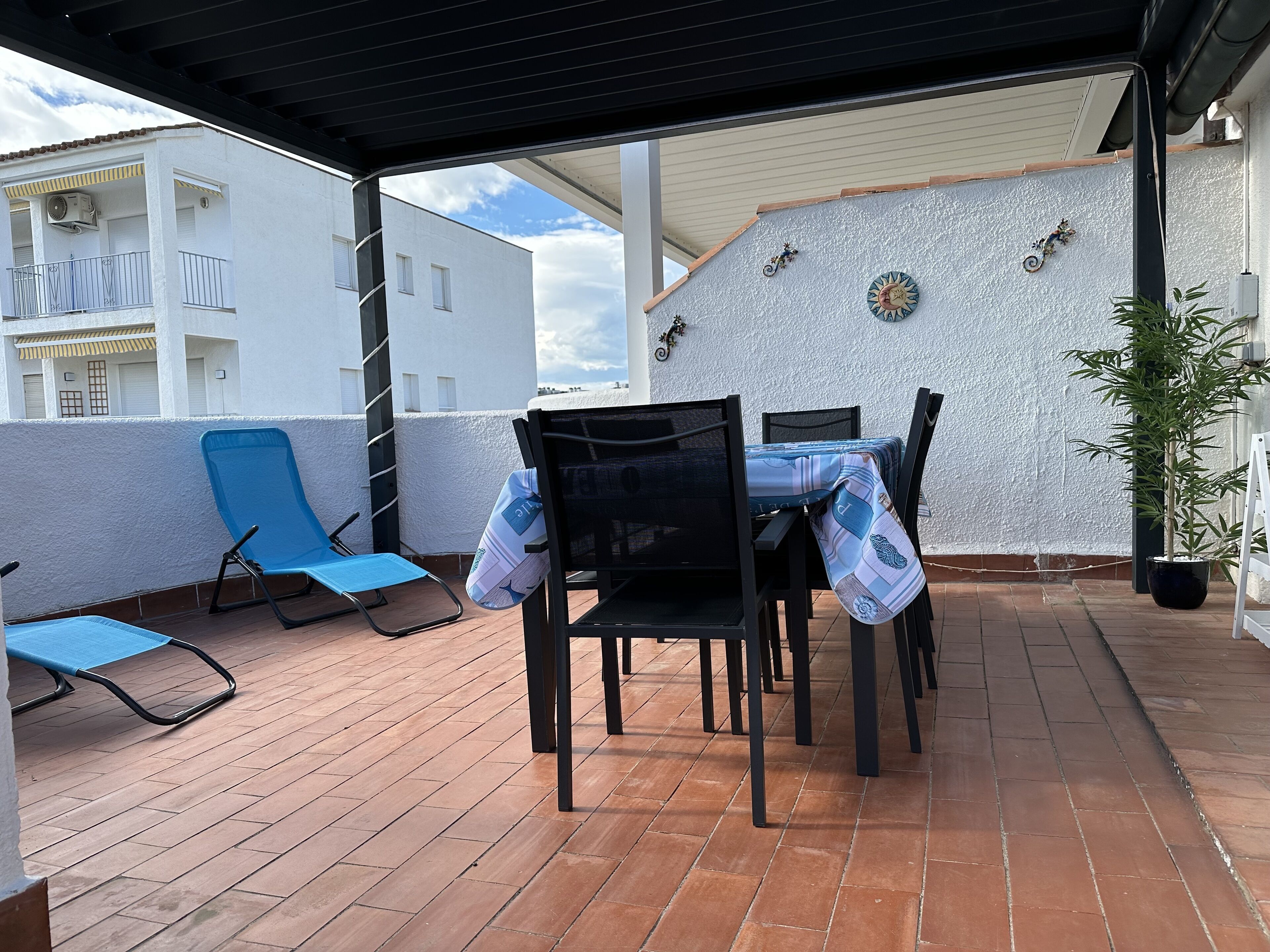 Un espace extérieur aménagé, abrité, avec une table et des chaises, un pot de fleurs et une vue sur un bâtiment blanc doté de balcons.
