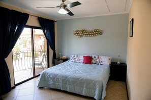 A bedroom with a bed, a ceiling fan, a nightstand, a bedside lamp, and a view through a window with curtains.