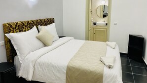 A neatly made bed with white linens and a beige throw blanket, a bedside table, a mirror above a sink, and a door with a light above it. - Dar Riad Jomana (Marrakech)