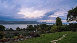 Ein Garten mit einem Kiesweg, der zu einem kleinen Haus am See führt, umgeben von viel Grün und Hügeln im Hintergrund.