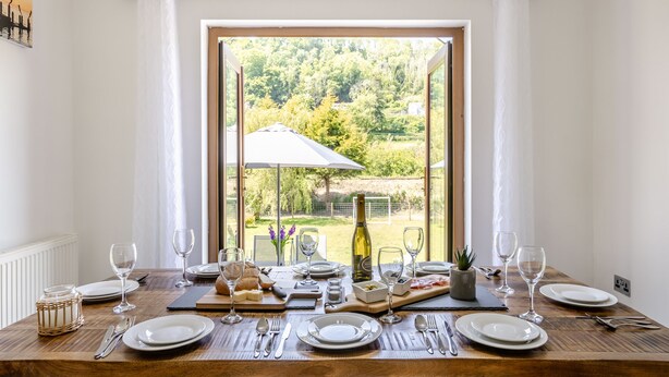 Une table à manger dressée pour six personnes, avec vue sur un jardin par une grande fenêtre.