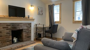 A living room with a fireplace, a sofa, a TV, and a desk with a chair. - New York Vermont Border Cottage Getaway (Granville)