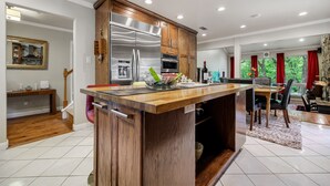 A modern kitchen with a central island, wooden cabinets, stainless steel appliances, and a dining area with a view of greenery.