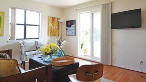 A living room with a sofa, a coffee table with a vase of flowers, a flat-screen TV mounted on the wall, and large windows with curtains.