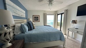 Quarto com cama, ventilador de teto, mesa de cabeceira, escrivaninha e janela com cortinas.