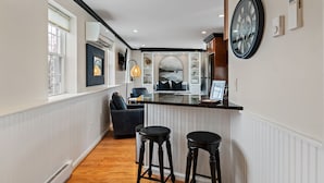 A narrow hallway with a kitchen area featuring a large clock, a refrigerator, and a seating area with two stools. - Cozy Loft for 2- Steps from Village of Pittsford (Pittsford)