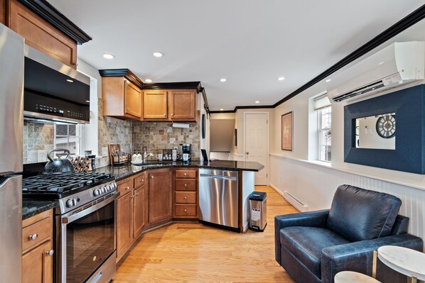 A modern kitchen with wooden cabinets, a stainless steel stove, and a built-in oven. There is a dining area with a blue leather sofa and a small round table. - Cozy Loft for 2- Steps from Village of Pittsford (Pittsford)