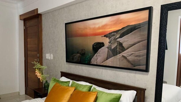 Un dormitorio con cama, almohadas, una mesita de noche, un jarrón con flores y un cuadro en la pared.