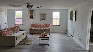A spacious living room with a beige sofa, a smaller sofa with floral cushions, a coffee table, and a ceiling fan. There are two windows providing natural light and a flat-screen TV mounted on the wall. - Family Ranch in pahrump (Pahrump)