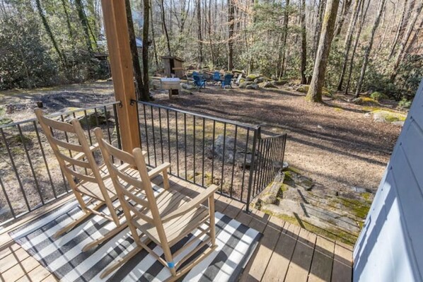 A wooden deck with a checkered mat, a metal railing, and a view of a forested area with a picnic table and chairs. - Ole Blue - Table Rock, Sassafras, Lake Keowee, Lake Jocassee. (Sunset)