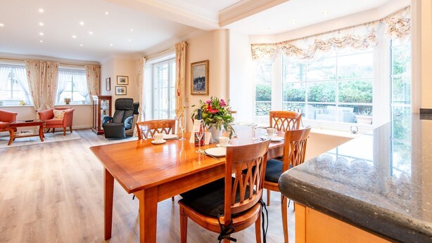 A spacious dining area with a wooden table and chairs, large windows with curtains, and a view of greenery outside. - House Lucia, Whg. 01 - House Lucia (Sylt)