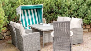 A wicker outdoor seating area with a table and chairs, set against a backdrop of green foliage and a stone wall. - House Lucia, Whg. 01 - House Lucia (Sylt)