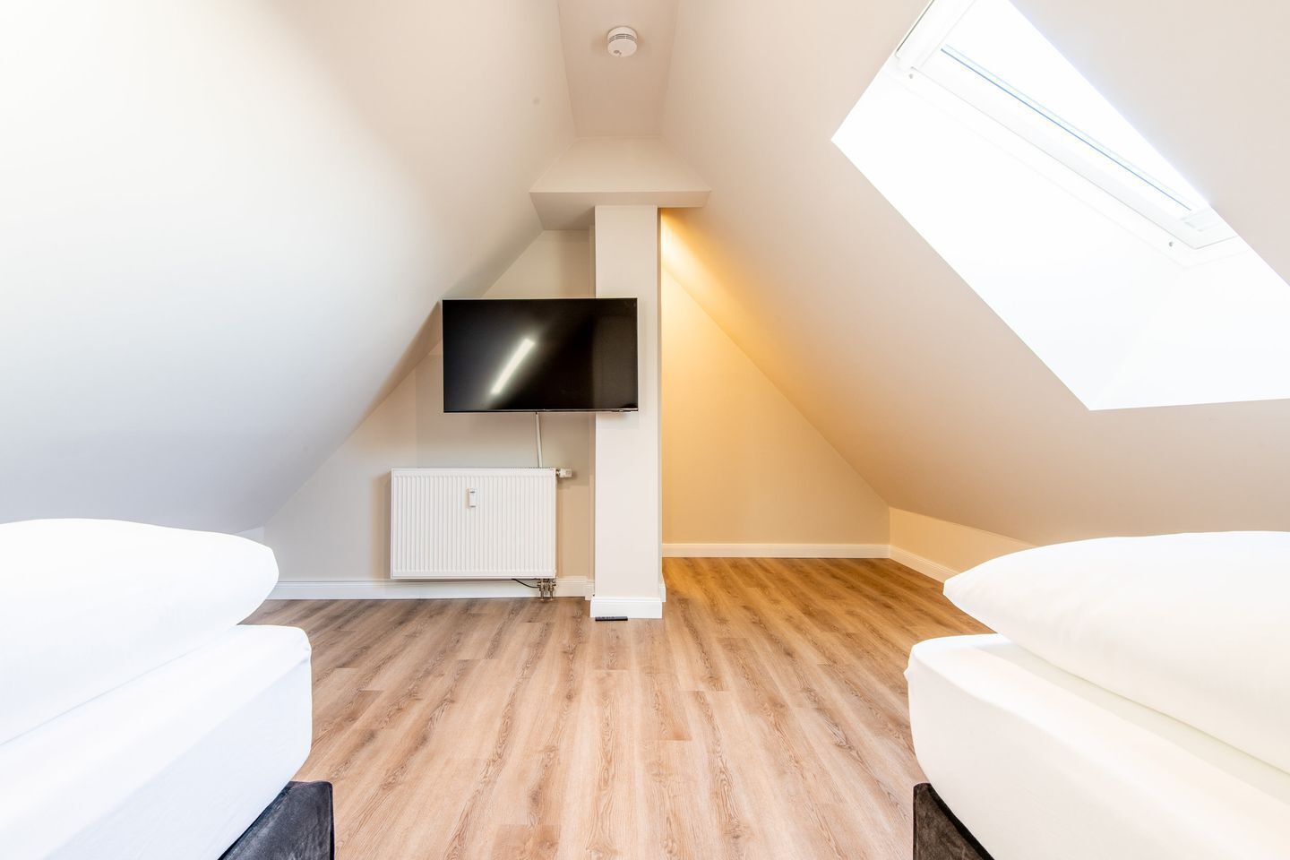 Una camera moderna e luminosa con soffitto spiovente, una TV a schermo piatto montata a parete, un termosifone bianco e pavimento in legno.