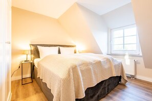 A bedroom with a sloped ceiling, a bed with a dark headboard, two bedside lamps, and a window with a view of greenery. - House Lucia, Whg. 04 - House Lucia (Sylt)