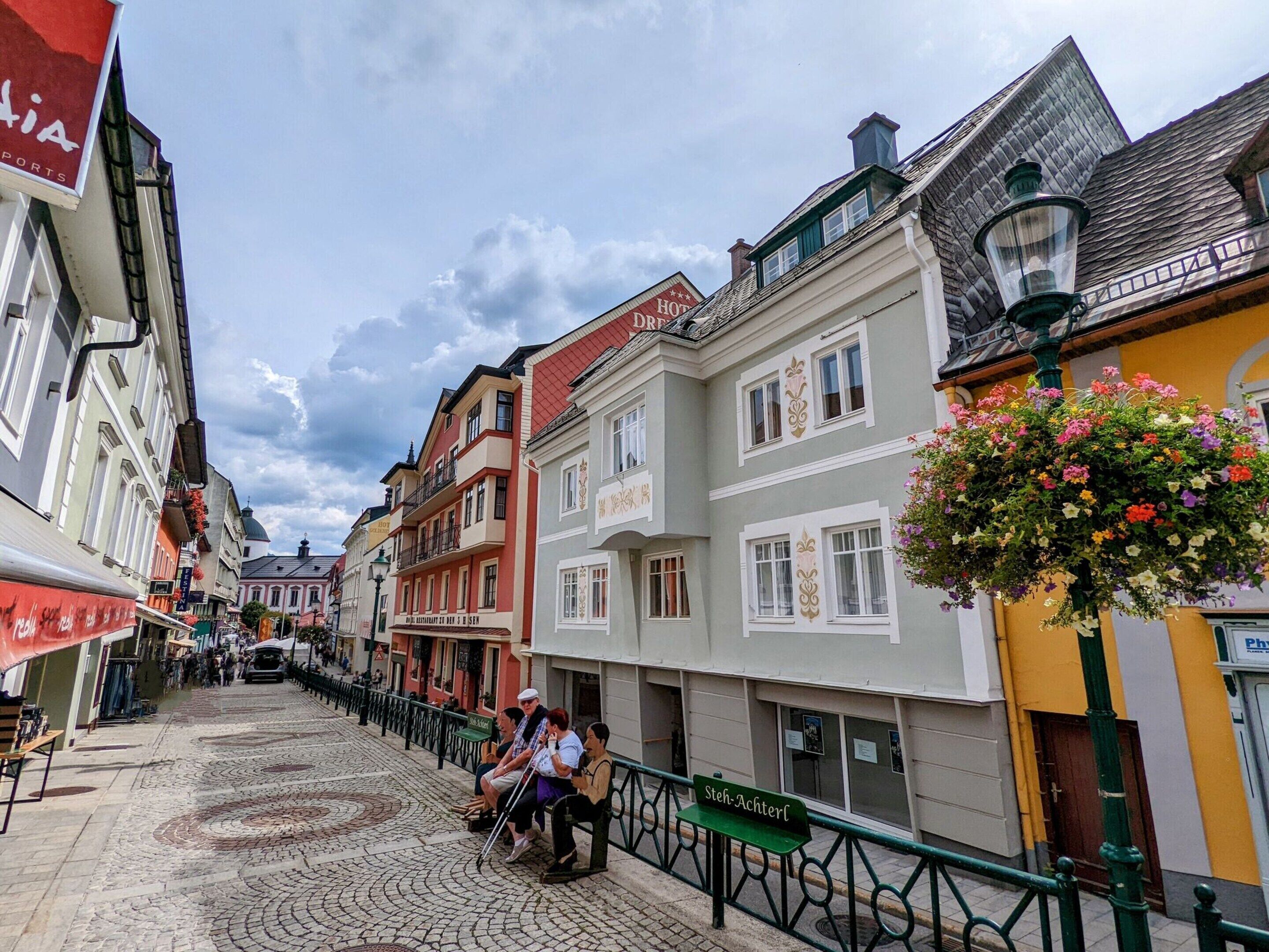 A cobblestone street lined with colorful buildings, a bench with people sitting, and a sign for 'Steh-Achterl'.