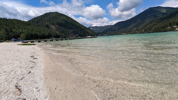 A beach with clear water, a wooden pier, and forested hills in the background. - Apartment in Mariazell Near Ski Slopes (Mariazell)