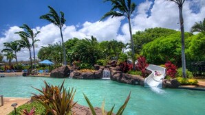 A pool with a water slide, surrounded by tropical plants and palm trees. - NEW! Top Floor Views with A/C in Poipu Beach, Resort Amenities (Koloa)