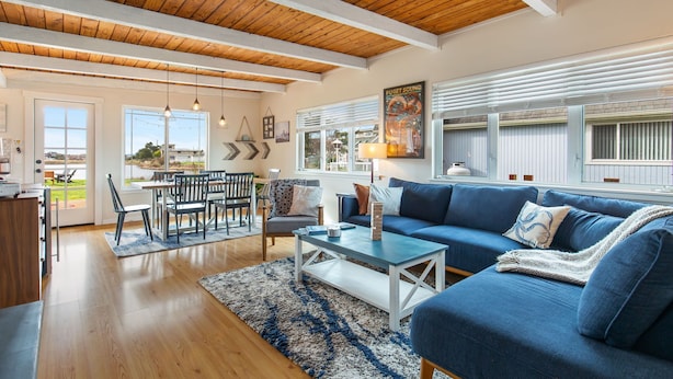 Ein geräumiges Wohnzimmer mit Holzbalkendecke, einem blauen Sofa, einem Essbereich mit Tisch und Stühlen sowie großen Fenstern mit Jalousien.