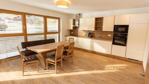 A modern kitchen with wooden cabinets, a dining table with chairs, and a large window with blinds. - Spacious Family Flat in Kleinarl (Kleinarl)