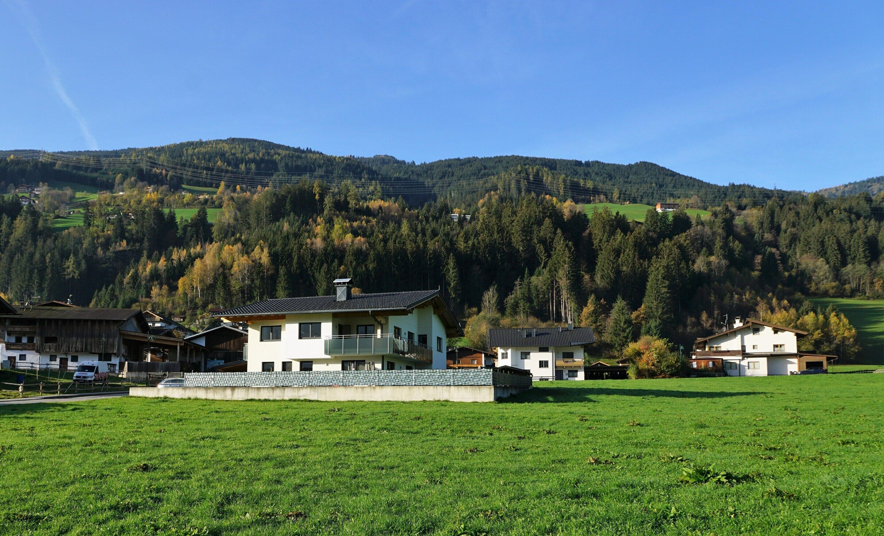 A rural setting with a large white building with a balcony, surrounded by green fields and forested hills.