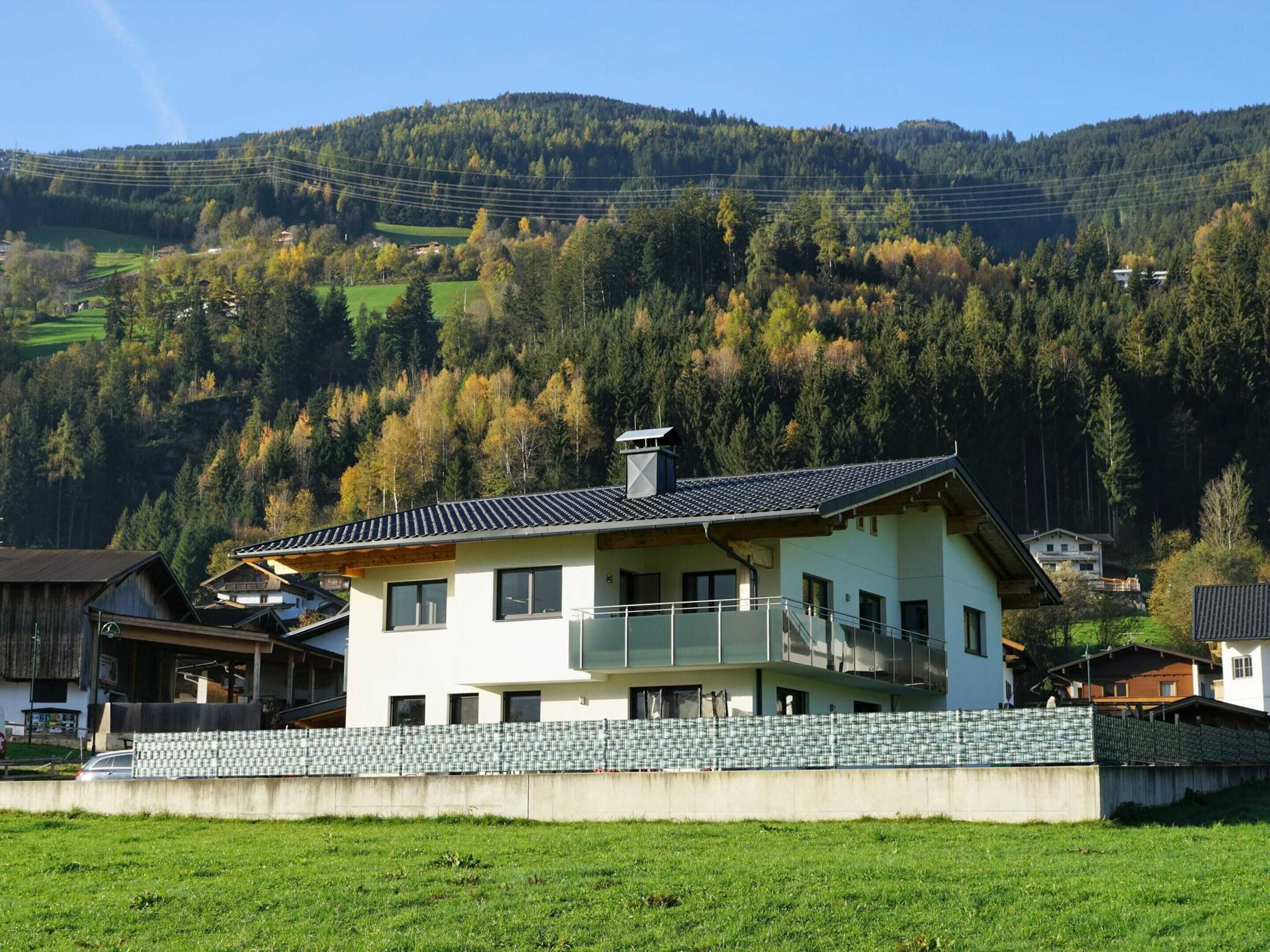 A two-story house with a balcony, surrounded by a stone wall, set against a backdrop of a forested hillside.