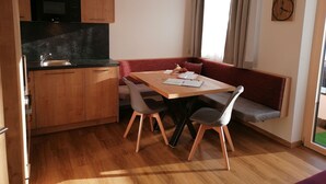 A compact kitchen with wooden cabinets, a black countertop, and a built-in microwave. A dining table with two chairs and a red bench is positioned near a window with curtains. - Contemporary Apartment in Gerlosberg Near Forest (Gerlosberg)