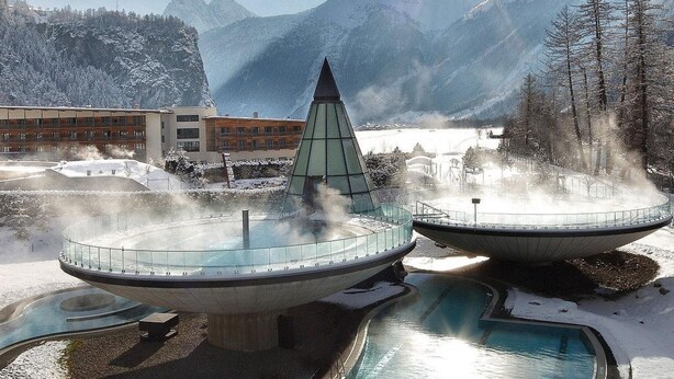 Un espace de bien-être doté de deux bains à remous entourés de neige, avec une vue sur les montagnes et un bâtiment aux nombreuses fenêtres.