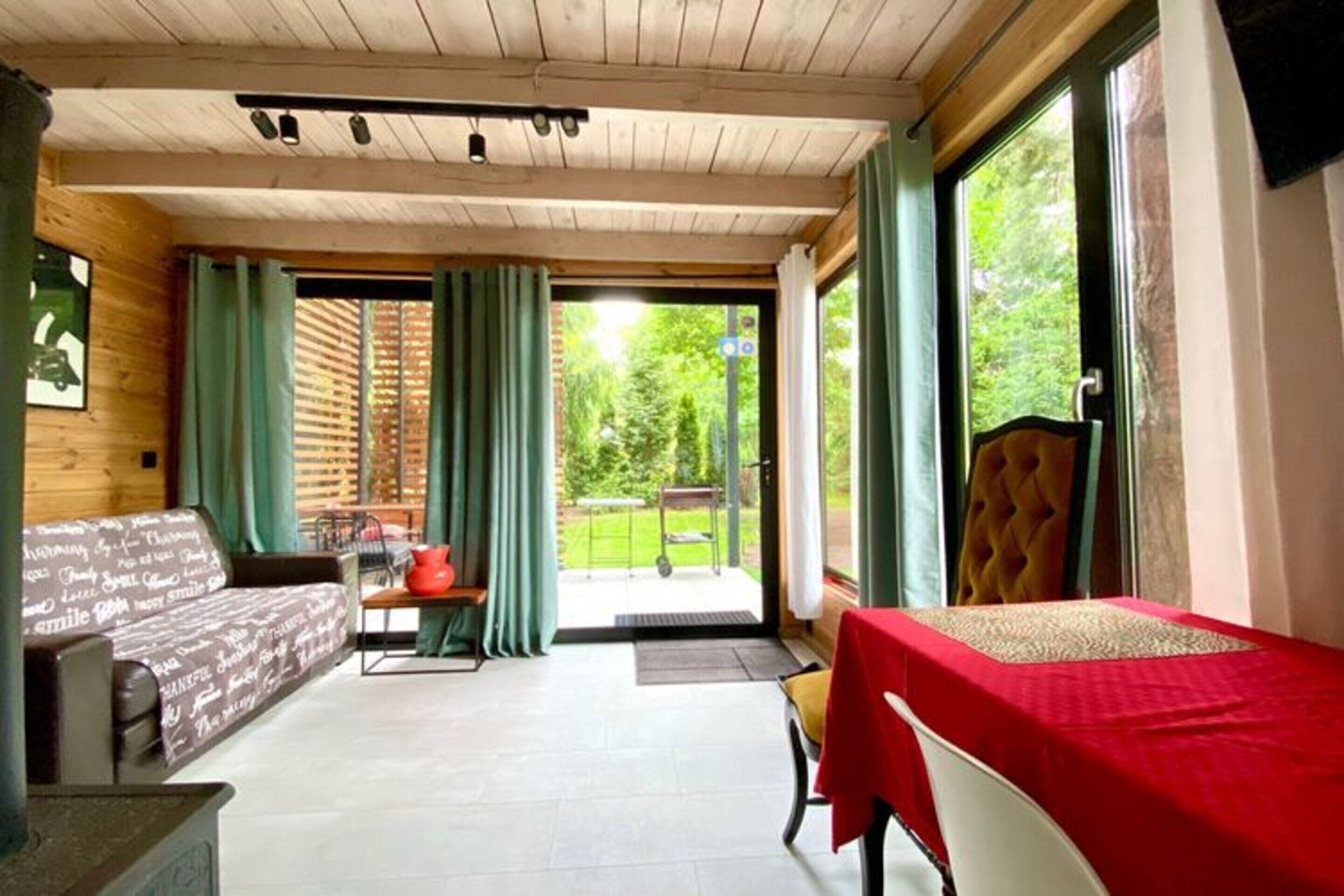 A cozy room with a sofa, a table with a red tablecloth, and a bed with a red bedspread. There are curtains on the windows, and a view of the outdoors is visible.
