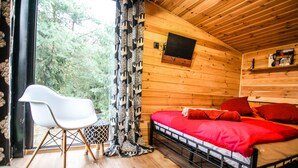 Una habitación de cabaña de madera con una cama, una silla, un televisor y un ventanal con cortinas.