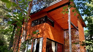 Una cabaña moderna de madera con ventanas amplias, un patio con mobiliario exterior y un bosque circundante.
